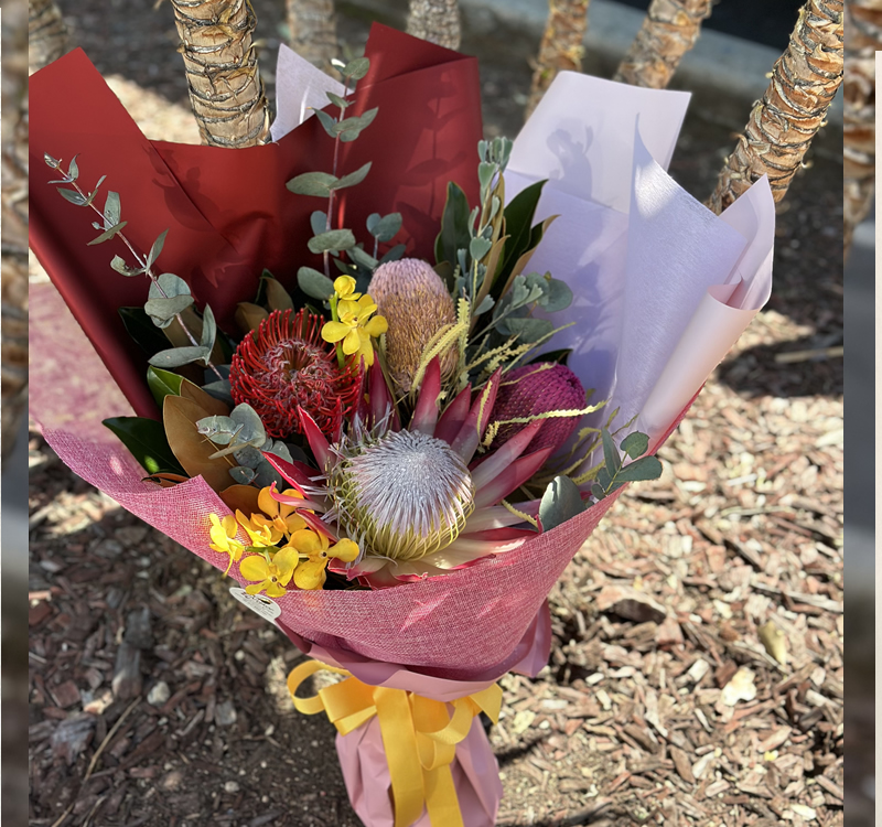 A colorful bouquet of flowers wrapped in maroon, white, and pink paper, tied with a yellow ribbon, placed on a mulched ground with the base of several tree trunks in the background.