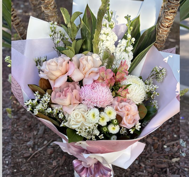 A bouquet of pink roses, white chrysanthemums, and assorted flowers wrapped in light purple and white paper with a purple bow at the base.