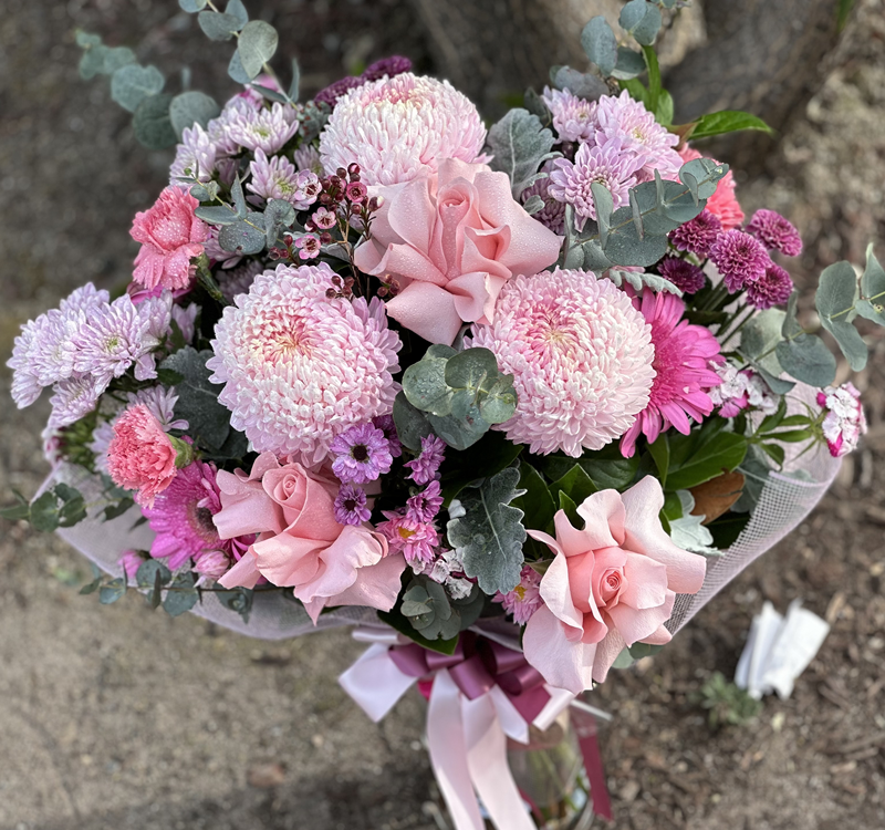 A bouquet of pink and purple flowers, including roses, chrysanthemums, and eucalyptus leaves, arranged with a pink ribbon and wrapping, placed outdoors.